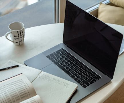 A cozy indoor workspace featuring a laptop, open book, notepad, and coffee, perfect for studying or remote work.