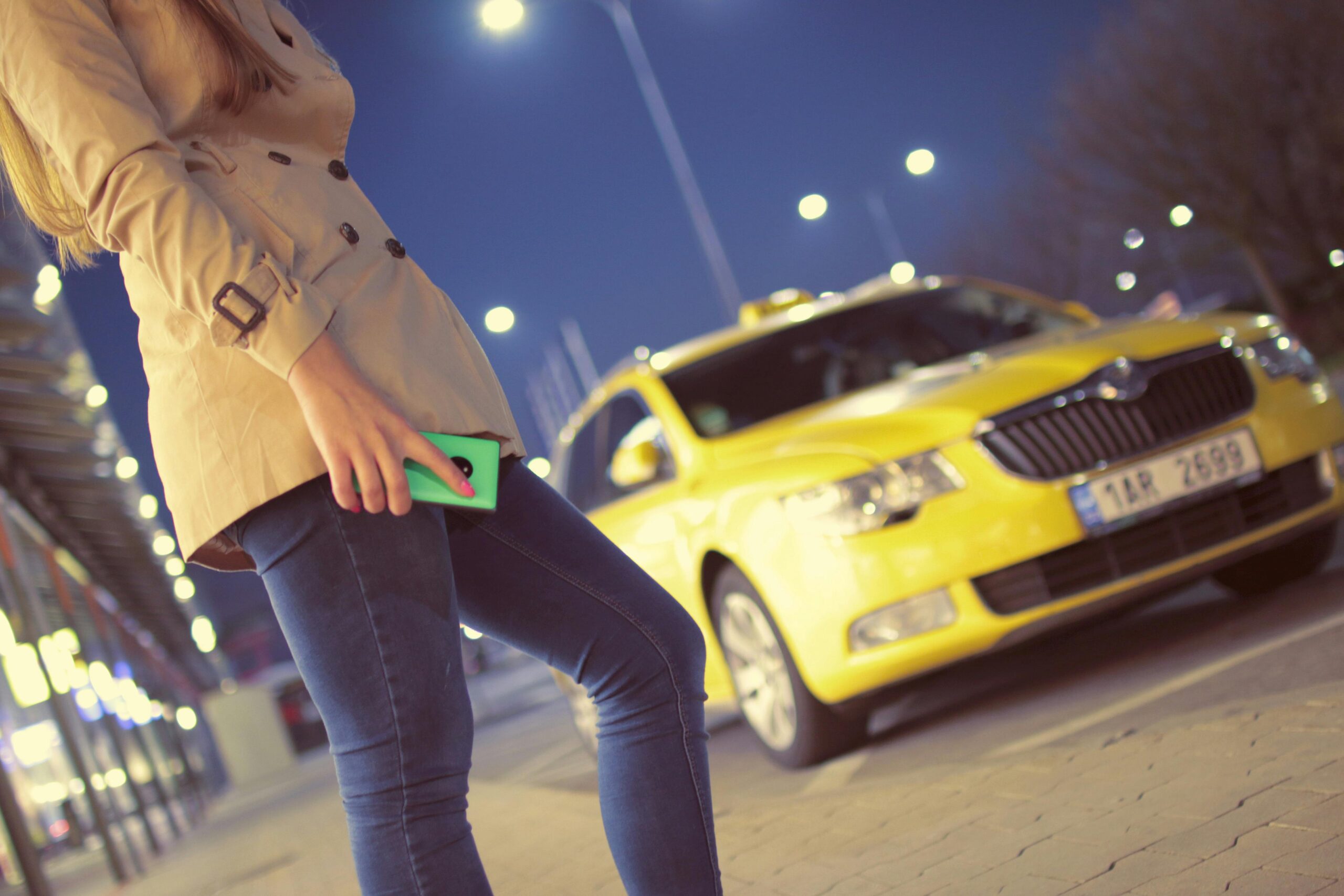 A woman in a coat stands with a smartphone by a yellow taxi under street lights at night.